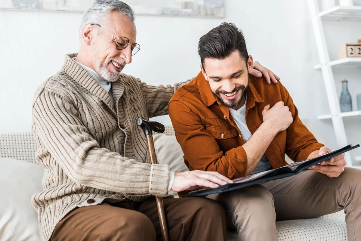 A senior man with a walking cane puts his arms around his son as they sit and look at family photos