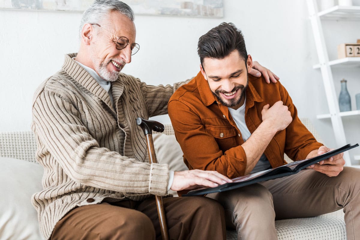 A senior man with a walking cane puts his arms around his son as they sit and look at family photos