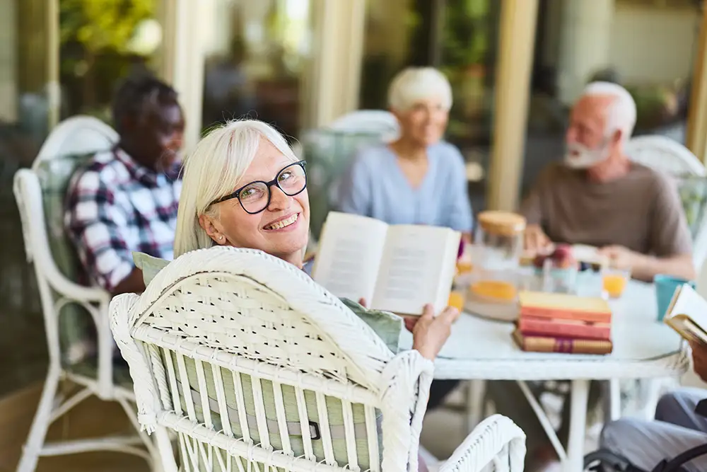 A smiling senior woman with glasses holds an open book while sitting outdoors with a group of friends around a patio table, highlighting how The Heritage at Brentwood residents enjoy making friends after 60 through shared interests.