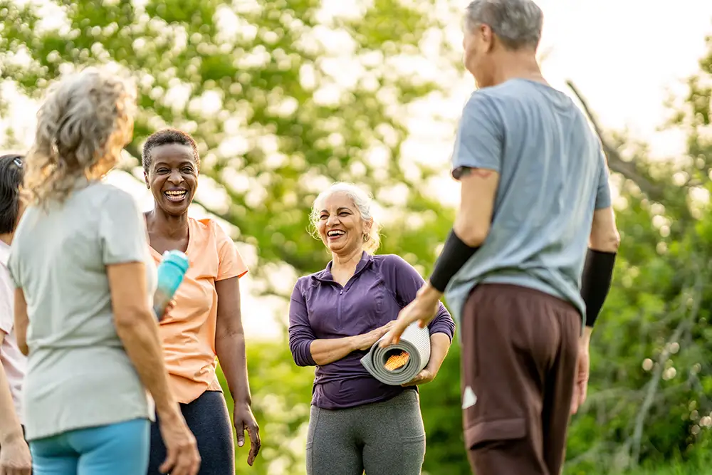 A group of active seniors at The Heritage at Brentwood share a laugh outdoors in athletic wear while holding rolled yoga mats and water bottles, demonstrating how easy making friends after 60 can be through group exercise.