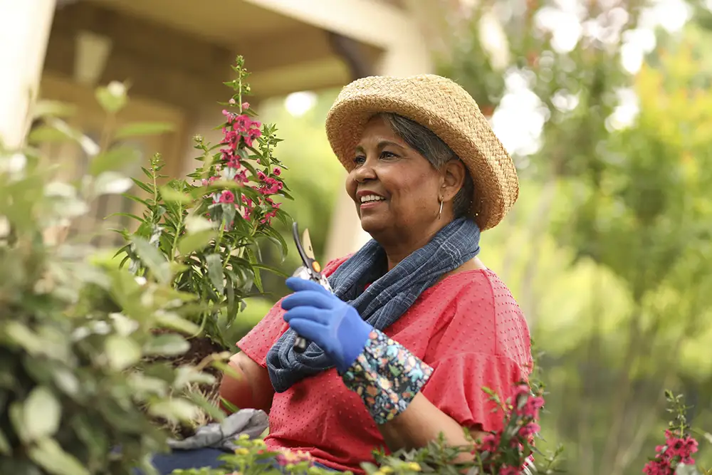 A senior woman in a straw hat smiles as she prunes flowers, enjoying gardening as part of her retirement bucket list at The Heritage at Brentwood®.