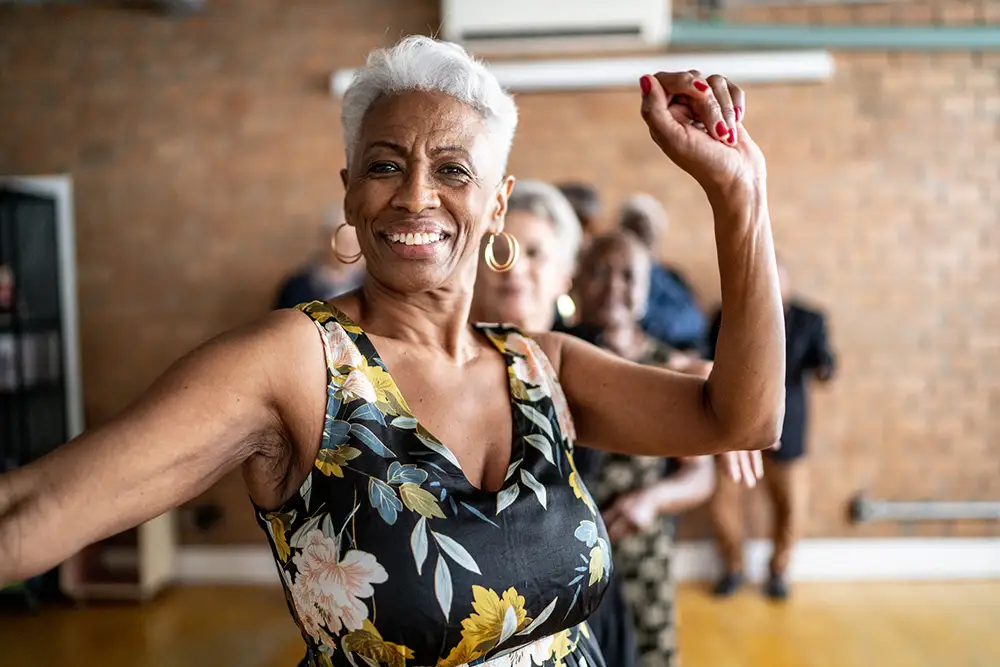 A smiling senior woman with short silver hair dances with her arm raised, fulfilling a item on her retirement bucket list alongside other seniors at The Heritage at Brentwood®.